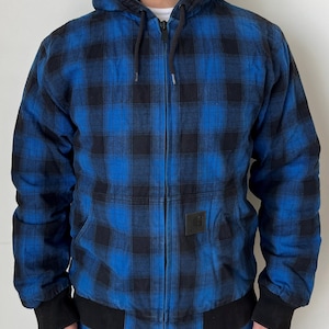 May include: Blue and black plaid hooded jacket with a full-length zipper. The jacket has a black hood lining, black cuffs, and a black bottom hem. A small square black patch is on the front. The jacket is worn over a white shirt.