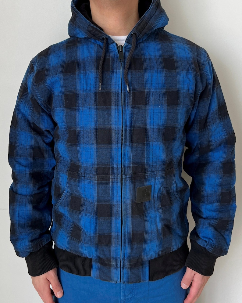 May include: Blue and black plaid hooded jacket with a full-length zipper. The jacket has a black hood lining, black cuffs, and a black bottom hem. A small square black patch is on the front. The jacket is worn over a white shirt.