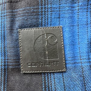 May include: Close-up of a blue and black plaid fabric with a black leather patch. The patch features the Carhartt logo and brand name in white lettering. The fabric has a woven texture.