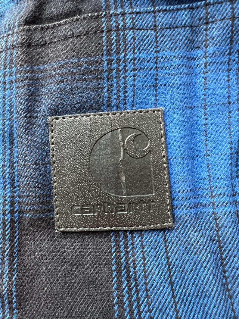 May include: Close-up of a blue and black plaid fabric with a black leather patch. The patch features the Carhartt logo and brand name in white lettering. The fabric has a woven texture.