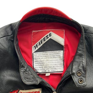 May include: Black leather motorcycle jacket with a red collar lining. Features the "DAINESE" logo on the chest and a label with text. Includes a zipper closure and snap details. Size 50.