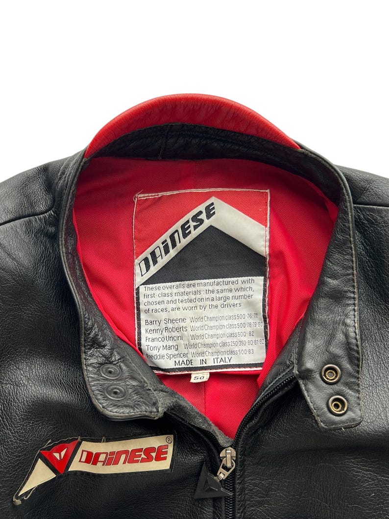 May include: Black leather motorcycle jacket with a red collar lining. Features the "DAINESE" logo on the chest and a label with text. Includes a zipper closure and snap details. Size 50.