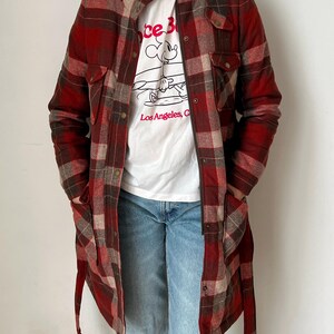 Op de afbeelding: Een lange, rood-bruine geruite jas met een knoopsluiting en zakken. Gedragen over een wit t-shirt met een Mickey Mouse-afbeelding en de tekst "Los Angeles, CA". De persoon draagt ook een blauwe spijkerbroek.