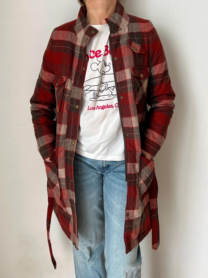 Op de afbeelding: Een lange, rood-bruine geruite jas met een knoopsluiting en zakken. Gedragen over een wit t-shirt met een Mickey Mouse-afbeelding en de tekst "Los Angeles, CA". De persoon draagt ook een blauwe spijkerbroek.
