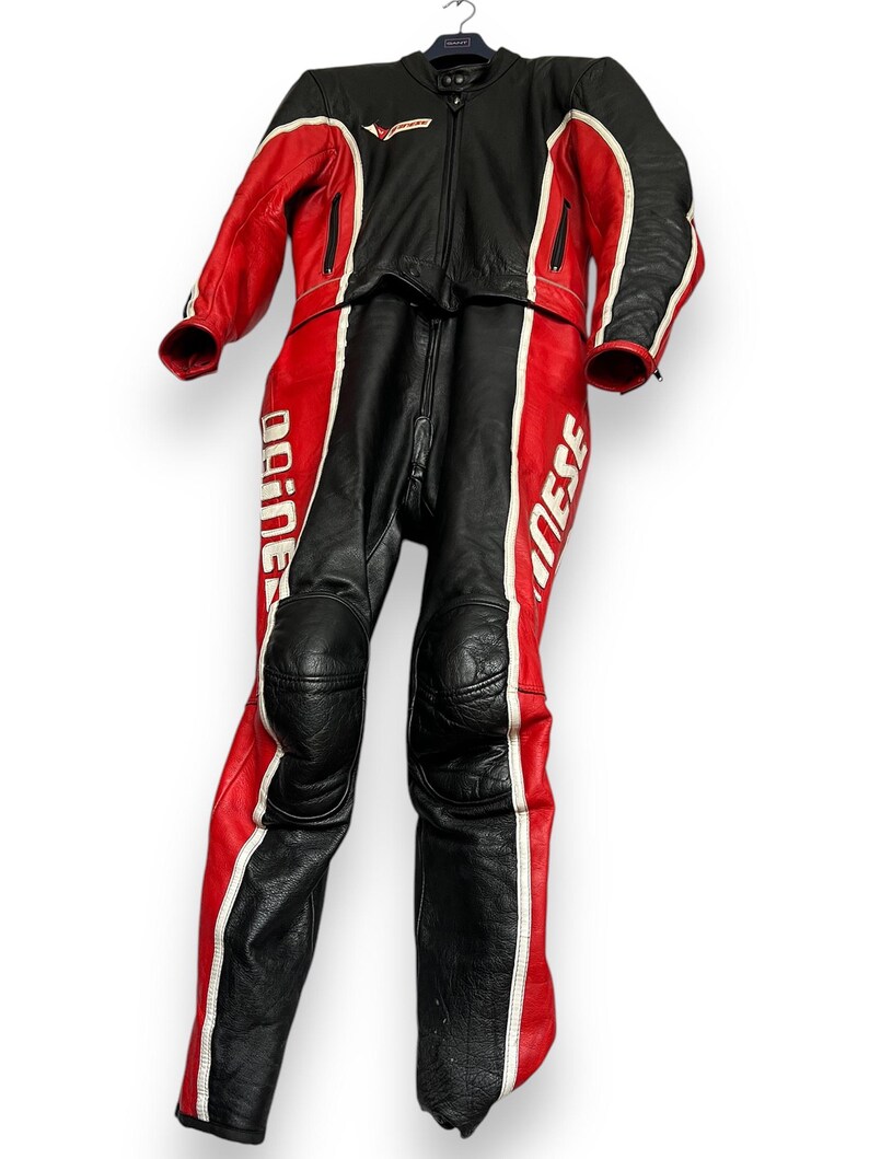 May include: Black and red motorcycle racing suit with white accents. The suit is made of leather and features a full-length zipper, knee pads, and the brand name "Dainese" printed on the legs. The suit is hanging on a hanger.