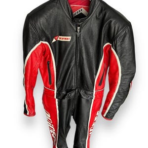 May include: Black and red leather motorcycle suit with white accents. The suit features a full-length zipper, multiple pockets, and the "Dainese" logo. The suit is hanging on a black hanger.