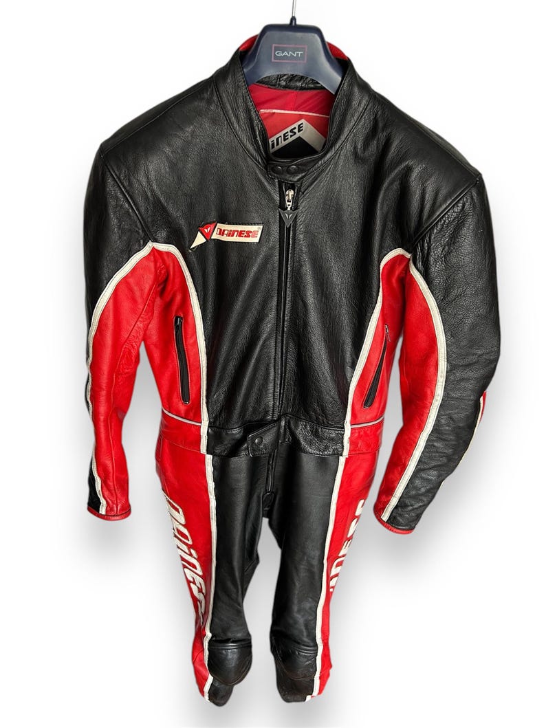 May include: Black and red leather motorcycle suit with white accents. The suit features a full-length zipper, multiple pockets, and the "Dainese" logo. The suit is hanging on a black hanger.