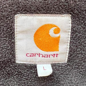 May include: Close-up of a Carhartt label, featuring the brand's logo in orange and white, with the brand name in pink. The label is on a textured, dark gray fabric. A small white tag with the letter "L" is attached.