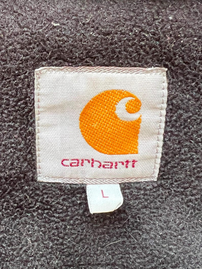 May include: Close-up of a Carhartt label, featuring the brand's logo in orange and white, with the brand name in pink. The label is on a textured, dark gray fabric. A small white tag with the letter "L" is attached.
