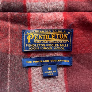 Op de afbeelding: Close-up van een rood en bordeaux geruite stof met twee blauwe labels. Het bovenste label luidt "PENDLETON" en "100% Virgin Wool". Het onderste label luidt "THE PORTLAND COLLECTION" en "MADE IN USA."