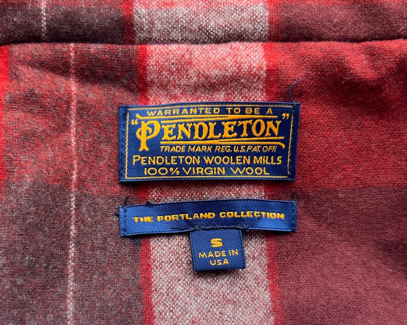 Op de afbeelding: Close-up van een rood en bordeaux geruite stof met twee blauwe labels. Het bovenste label luidt "PENDLETON" en "100% Virgin Wool". Het onderste label luidt "THE PORTLAND COLLECTION" en "MADE IN USA."