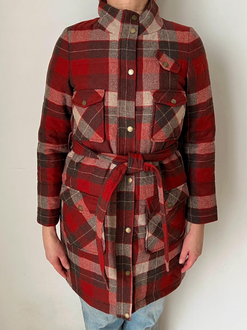 Op de afbeelding: Een rood, bruin en beige geruit jack met een ceintuur in de taille en vier voorzakken. De jas heeft een hoge kraag en wordt gesloten met goudkleurige knopen. De mouwen zijn lang en de jas is gemaakt van warm, geweven materiaal.