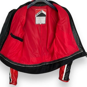 May include: Black leather motorcycle jacket with a red interior lining. The jacket has a zipper closure, a small black pocket, and red and white stripes on the sleeves. The interior label reads "Dainese".