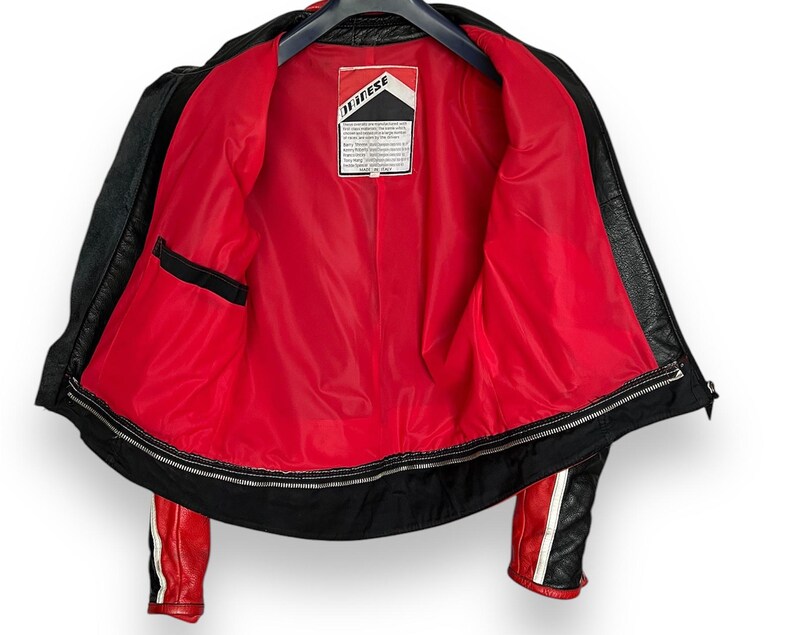 May include: Black leather motorcycle jacket with a red interior lining. The jacket has a zipper closure, a small black pocket, and red and white stripes on the sleeves. The interior label reads "Dainese".