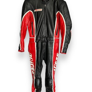 May include: Black and red leather motorcycle racing suit with white accents. The suit features a full-body design with a zipper closure, knee pads, and the "Dainese" logo. The suit is hanging on a hanger.
