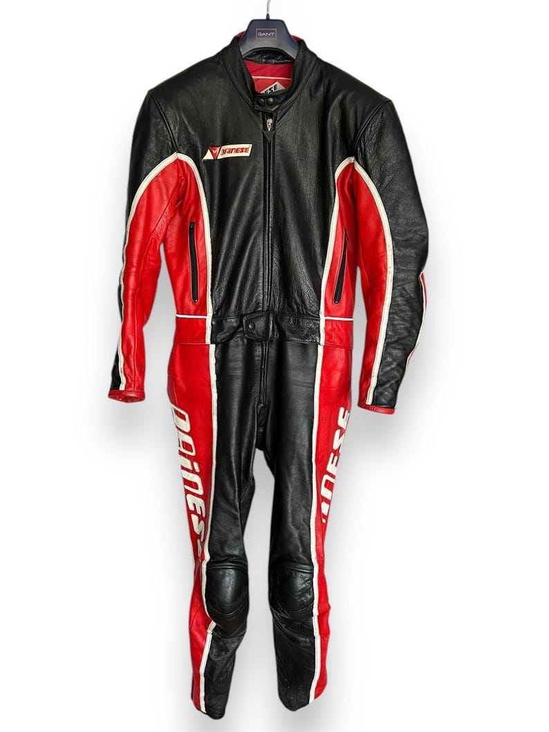 May include: Black and red leather motorcycle racing suit with white accents. The suit features a full-body design with a zipper closure, knee pads, and the "Dainese" logo. The suit is hanging on a hanger.