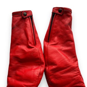 May include: Pair of bright red leather pants, featuring black zippers and button closures at the cuffs. The leather has a textured appearance, and the pants are set against a white background.