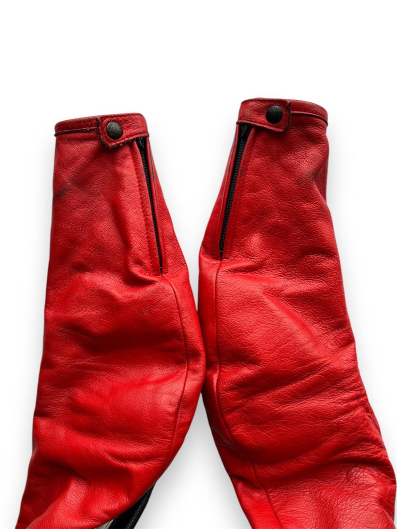 May include: Pair of bright red leather pants, featuring black zippers and button closures at the cuffs. The leather has a textured appearance, and the pants are set against a white background.
