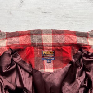 Op de afbeelding: Close-up van een rood, bruin en beige geruit jack. De jas heeft een bordeauxrode satijnen voering en een Pendleton-label. De kraag heeft twee knopen en een rits. De jas maakt deel uit van The Portland Collection.