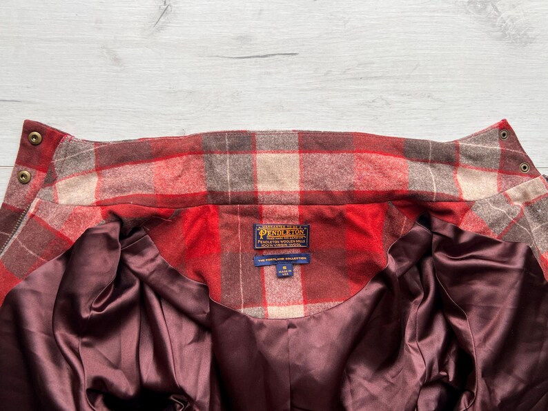 Op de afbeelding: Close-up van een rood, bruin en beige geruit jack. De jas heeft een bordeauxrode satijnen voering en een Pendleton-label. De kraag heeft twee knopen en een rits. De jas maakt deel uit van The Portland Collection.
