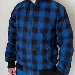 May include: Blue and black plaid hooded jacket with a zip-up front, black ribbed cuffs, and a black ribbed hem. The jacket has a square black patch on the front and a drawstring hood. The jacket is worn over a white shirt.