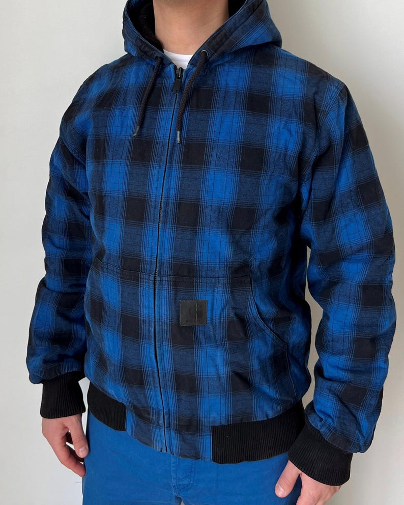 May include: Blue and black plaid hooded jacket with a zip-up front, black ribbed cuffs, and a black ribbed hem. The jacket has a square black patch on the front and a drawstring hood. The jacket is worn over a white shirt.