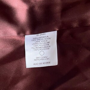 Op de afbeelding: Close-up van een bordeauxrode stof met een wit label. Het label geeft aan dat de buitenkant 100% scheerwol is, de voering 100% polyester en de vulling 100% polyester. Het label zegt ook "Chemisch reinigen".