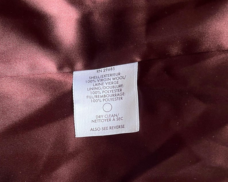 Op de afbeelding: Close-up van een bordeauxrode stof met een wit label. Het label geeft aan dat de buitenkant 100% scheerwol is, de voering 100% polyester en de vulling 100% polyester. Het label zegt ook "Chemisch reinigen".