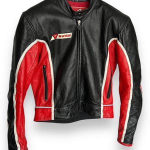 May include: Black leather motorcycle jacket with red and white accents. Features a zip-up front, zippered pockets, and the "Dainese" logo on the chest. The jacket is hanging on a black hanger.
