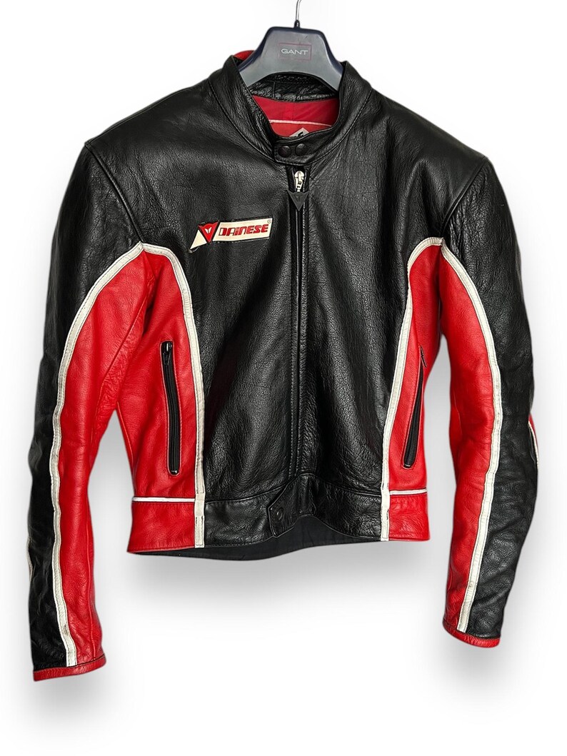May include: Black leather motorcycle jacket with red and white accents. Features a zip-up front, zippered pockets, and the "Dainese" logo on the chest. The jacket is hanging on a black hanger.