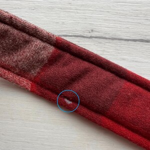 Op de afbeelding: Close-up van een rood en bordeaux geruite hoofdband. De hoofdband is gemaakt van een zachte, getextureerde stof. Het ruitpatroon heeft een mix van rode en bordeaux tinten, wat een warme uitstraling cre&euml;ert. De hoofdband is ontworpen om om het hoofd te worden gedragen.