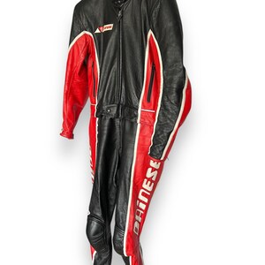 May include: Black and red leather motorcycle racing suit with white accents. The suit features a full-length zipper, knee pads, and the brand name "Dainese" printed on the leg. The suit is hanging on a hanger.