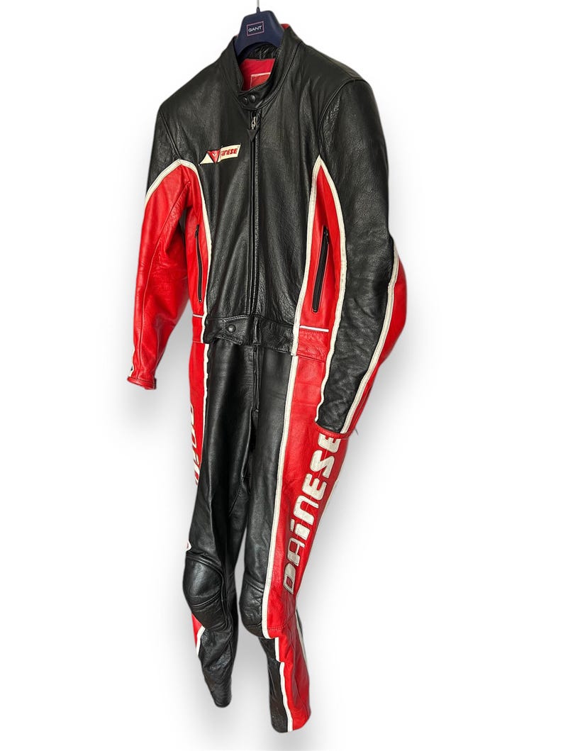 May include: Black and red leather motorcycle racing suit with white accents. The suit features a full-length zipper, knee pads, and the brand name "Dainese" printed on the leg. The suit is hanging on a hanger.