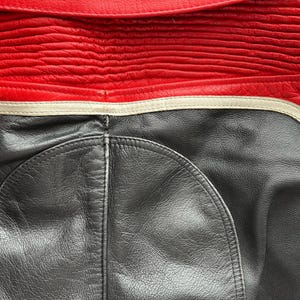 May include: Close-up of a red and black leather jacket. The top portion is red with horizontal stitched detailing. The bottom section is black leather with a curved design. A white stripe separates the two colors.