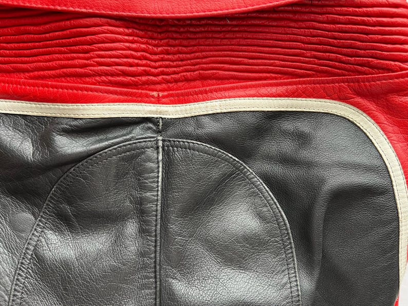 May include: Close-up of a red and black leather jacket. The top portion is red with horizontal stitched detailing. The bottom section is black leather with a curved design. A white stripe separates the two colors.