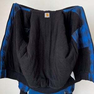 May include: Black zip-up jacket with blue plaid accents on the sleeves and shoulders. The interior is black, and a small Carhartt logo patch is visible. The jacket has a ribbed hem and cuffs.