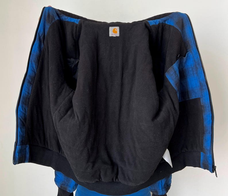 May include: Black zip-up jacket with blue plaid accents on the sleeves and shoulders. The interior is black, and a small Carhartt logo patch is visible. The jacket has a ribbed hem and cuffs.