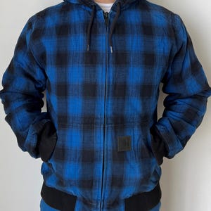 May include: A blue and black plaid hooded jacket with a full zipper closure. The jacket has black ribbed cuffs and a black ribbed hem. A small black square patch is on the front. The jacket is worn over a white shirt.