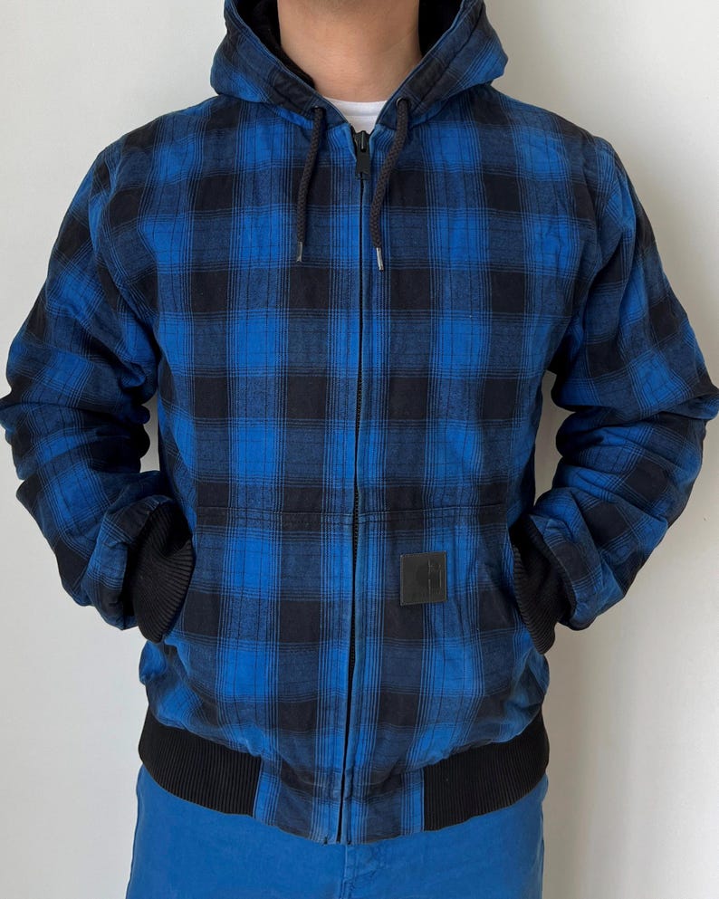 May include: A blue and black plaid hooded jacket with a full zipper closure. The jacket has black ribbed cuffs and a black ribbed hem. A small black square patch is on the front. The jacket is worn over a white shirt.