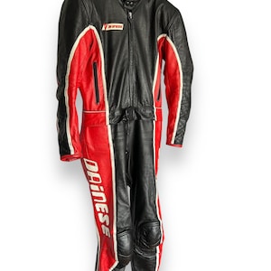 May include: Black and red leather motorcycle racing suit with white accents. The suit features the "Dainese" logo on the leg and a zipper closure. The suit is hanging on a hanger.