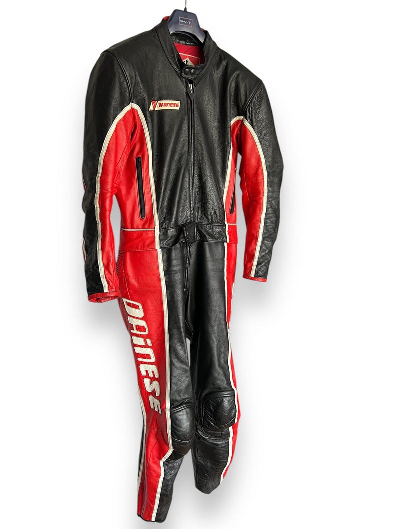 May include: Black and red leather motorcycle racing suit with white accents. The suit features the "Dainese" logo on the leg and a zipper closure. The suit is hanging on a hanger.