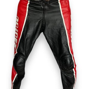 May include: Black leather motorcycle pants with red and white accents. The pants feature a zipper closure and knee padding. The word "Dainese" is printed in white on the red panels. The waist band has the word "Dainese" printed on it.