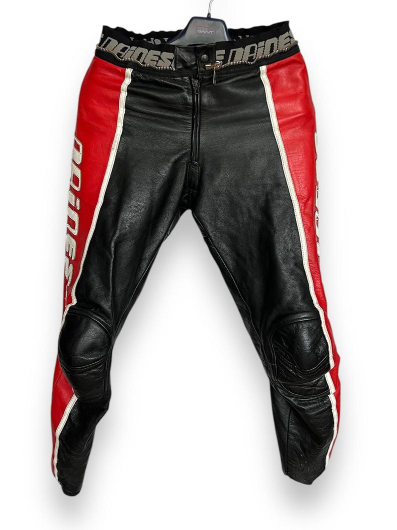 May include: Black leather motorcycle pants with red and white accents. The pants feature a zipper closure and knee padding. The word "Dainese" is printed in white on the red panels. The waist band has the word "Dainese" printed on it.