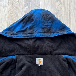 May include: Close-up of a blue and black plaid hooded jacket with a black fleece lining. The jacket has a Carhartt label. The hood is up, and the jacket is unzipped, revealing the interior.