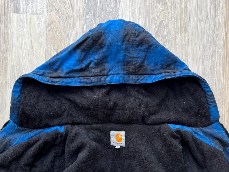 May include: Close-up of a blue and black plaid hooded jacket with a black fleece lining. The jacket has a Carhartt label. The hood is up, and the jacket is unzipped, revealing the interior.
