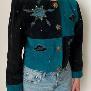 May include: Black and teal suede jacket with celestial embroidery. The jacket features a sun and cloud design with gold accents and decorative buttons. The sleeves and collar are trimmed in teal. A unique, vintage piece.