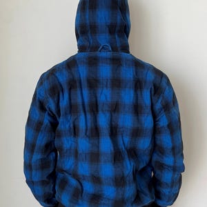 May include: A hooded jacket with a blue and black plaid pattern. The jacket has a black ribbed hem and cuffs. The hood is up, and the back of the jacket is visible against a white background. The jacket is a casual outerwear item.