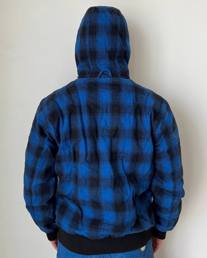 May include: A hooded jacket with a blue and black plaid pattern. The jacket has a black ribbed hem and cuffs. The hood is up, and the back of the jacket is visible against a white background. The jacket is a casual outerwear item.