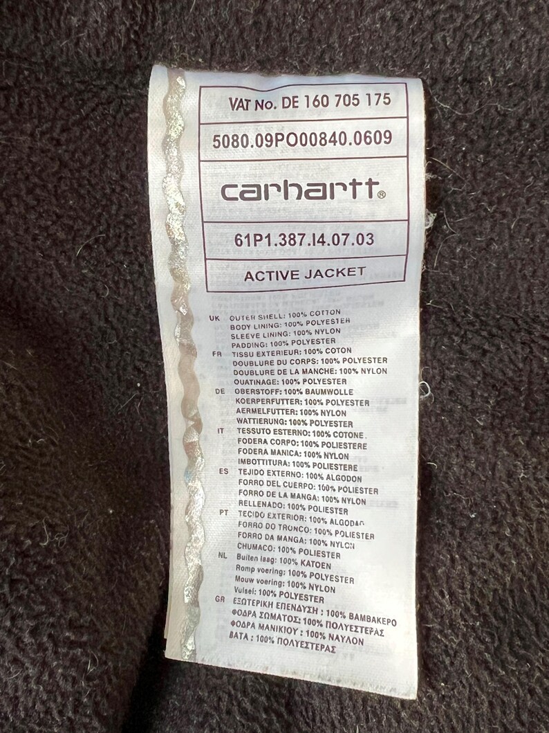 May include: A close-up of a Carhartt Active Jacket label. The label is white with black text, including the brand name and fabric composition details. The jacket's outer shell is 100% cotton, with a polyester body lining and nylon sleeve lining.