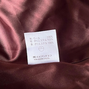 Op de afbeelding: Close-up van een bordeauxrode stoffen binnenkant met een wit label. Het label heeft Japanse tekst die de samenstelling van de stof aangeeft: 100% wol, 100% polyester, en een bedrijfsnaam en telefoonnummer.